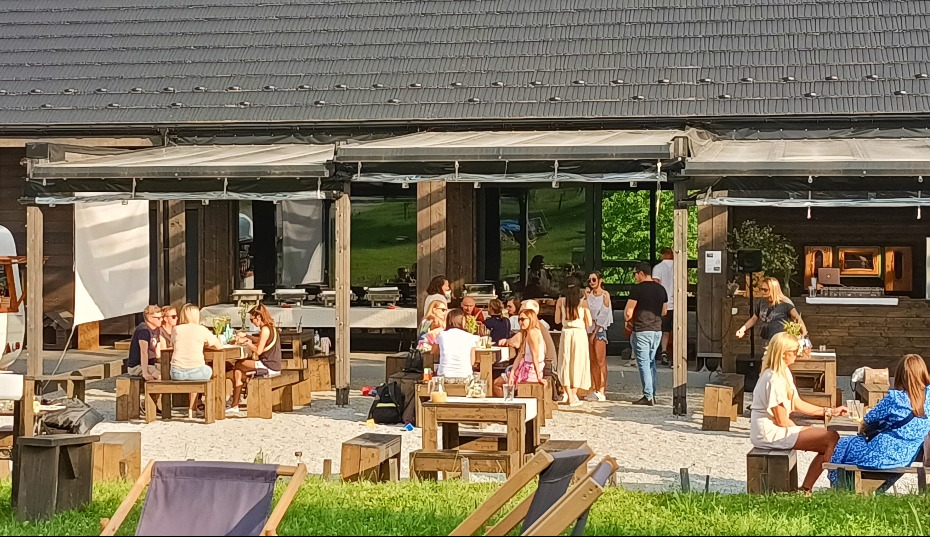 Zewnętrzna przestrzeń restauracji z drewnianymi meblami i gośćmi podczas pikniku; widoczne stoły z jedzeniem i napojami, bar z DJ-em w tle, oraz leżaki na pierwszym planie.