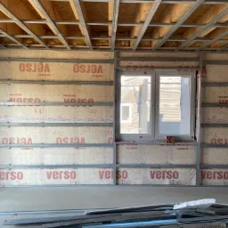 Wnętrze w trakcie montażu metalowej konstrukcji szkieletowej pod okładziny ścienne i sufitowe, widoczne profile, izolacja i folia paroizolacyjna z nadrukiem 'verso', okno w ścianie, na podłodze...