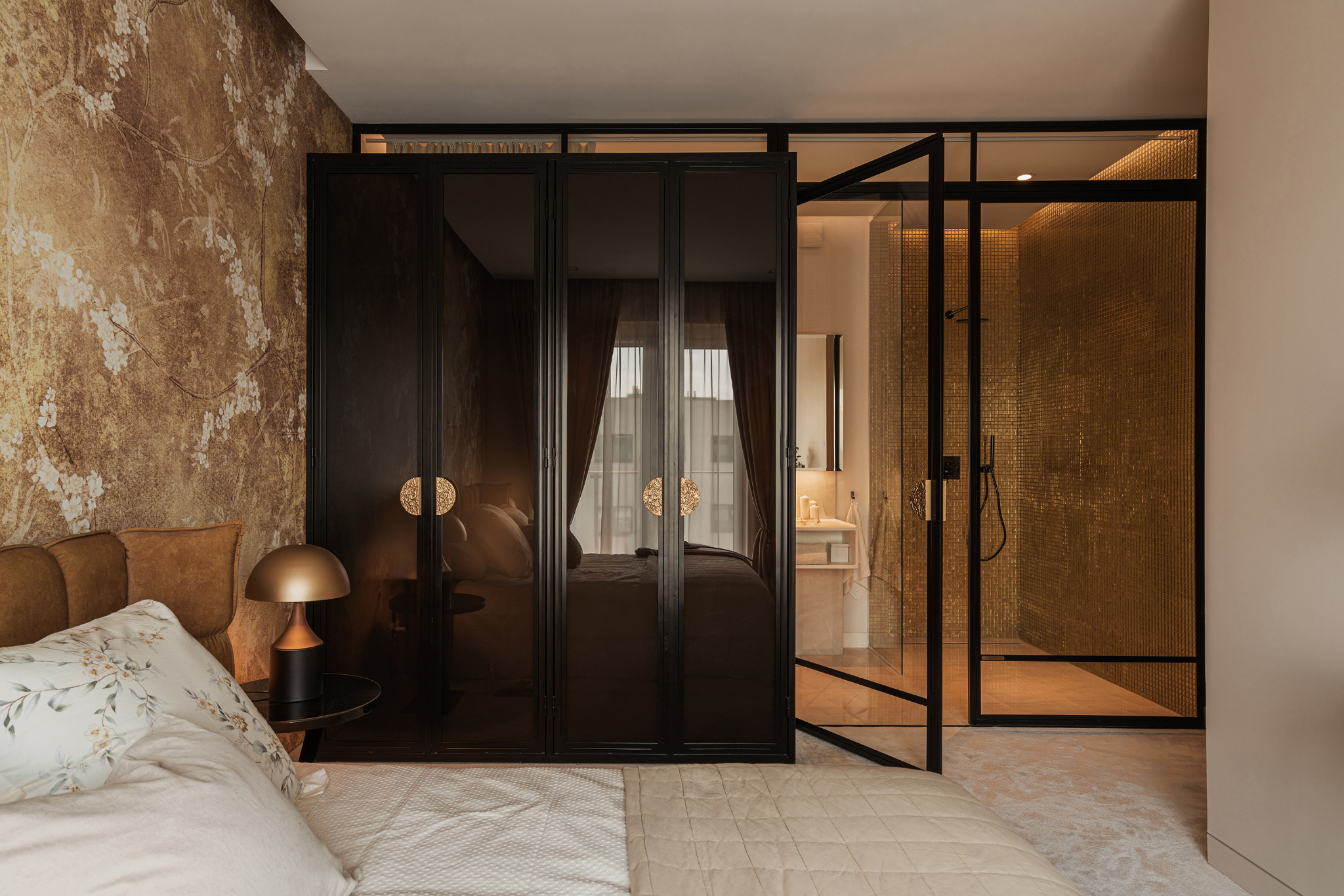 Sypialnia z łazienką en-suite: widok na łóżko z wzorzystą pościelą, szafę z ciemnego szkła z ozdobnymi uchwytami oraz przeszkloną kabinę prysznicową wyłożoną złotą mozaiką.