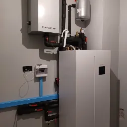 Kompletna instalacja grzewcza z pompą ciepła Midea, zbiornikiem wyrównawczym i orurowaniem w pomieszczeniu technicznym z szarą ścianą i rozdzielnicą elektryczną.