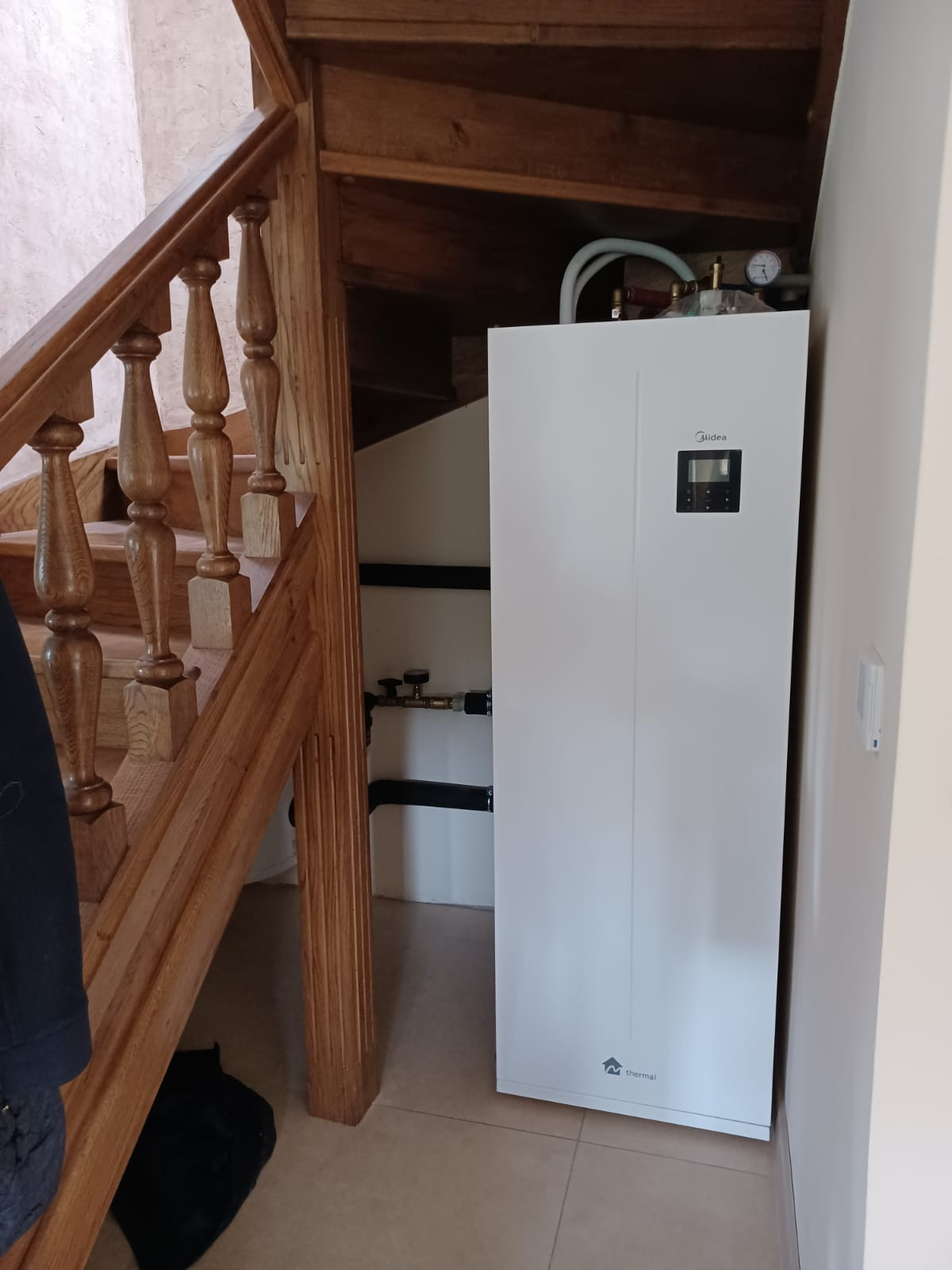 Biała pompa ciepła Midea Thermal zainstalowana w rogu pomieszczenia przy drewnianych schodach z balustradą, widoczne rury i manometr na górze urządzenia.