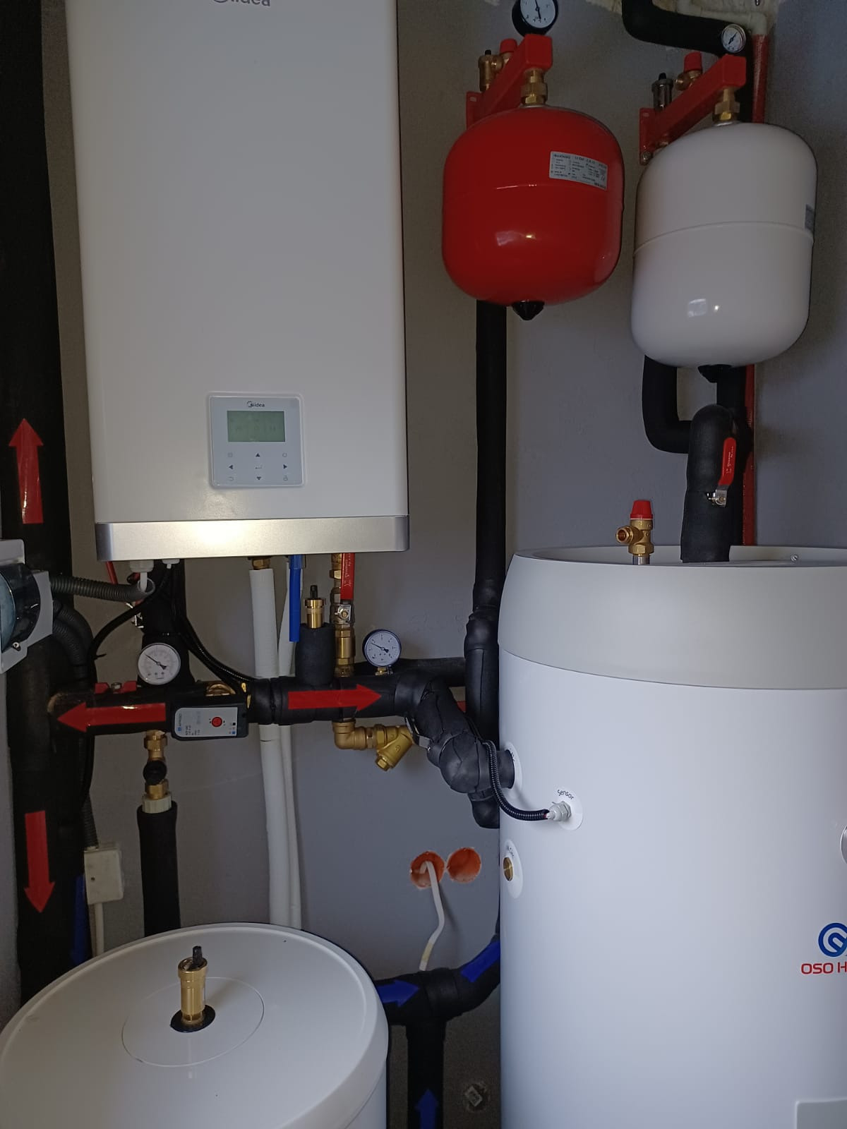 Instalacja grzewcza w kotłowni: biały piec Midea, czerwony i biały zbiornik wyrównawczy, biały zasobnik OSO Heating, liczne rury, zawory i manometry.