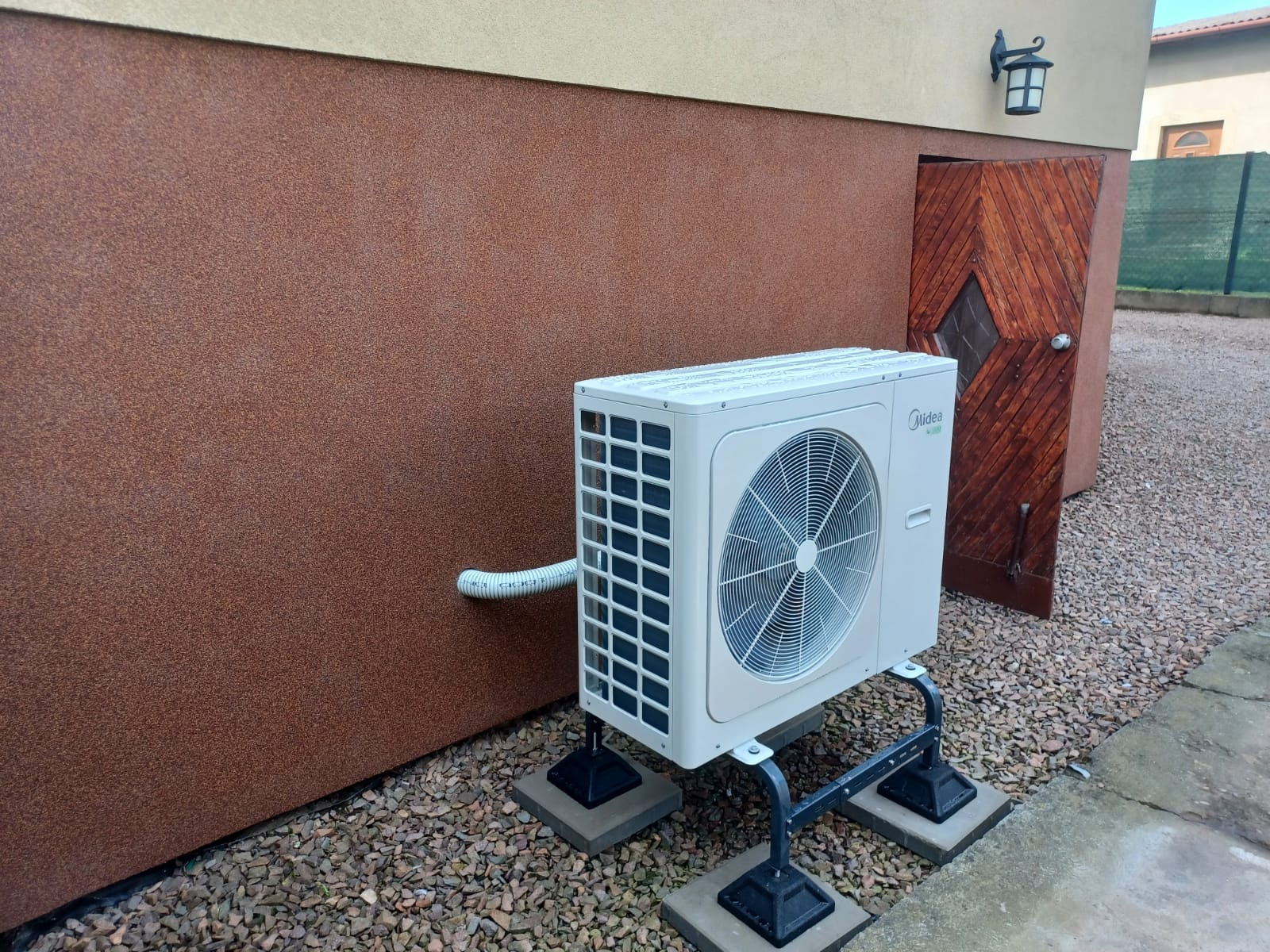 Zewnętrzna jednostka klimatyzacji Midea zamontowana na metalowej konstrukcji na betonowych podstawach, z brązową ścianą budynku w tle i drewnianymi drzwiami z lampionem nad nimi.