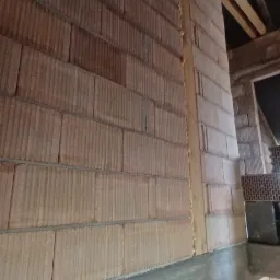 Wznoszenie ściany z bloczków ceramicznych na fundamencie z betonu, widoczne drewniane elementy konstrukcji dachu i pustaki wentylacyjne w tle.