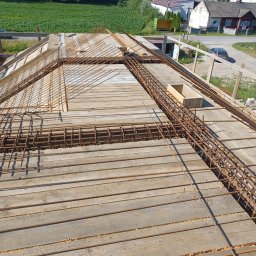 Uslugi budowlane Wacław smoter - Zbrojenie stropu na drewnianym szalunku, widoczne pręty zbrojeniowe i betonowa skrzynka, w tle zabudowa wiejska i pole uprawne.