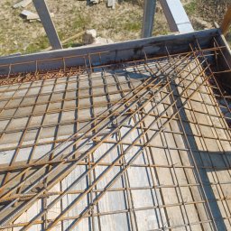Uslugi budowlane Wacław smoter - Zbrojenie fundamentu z siatki stalowej na deskowaniu, widoczne elementy konstrukcji szalunkowej i fragment gruntu w tle.