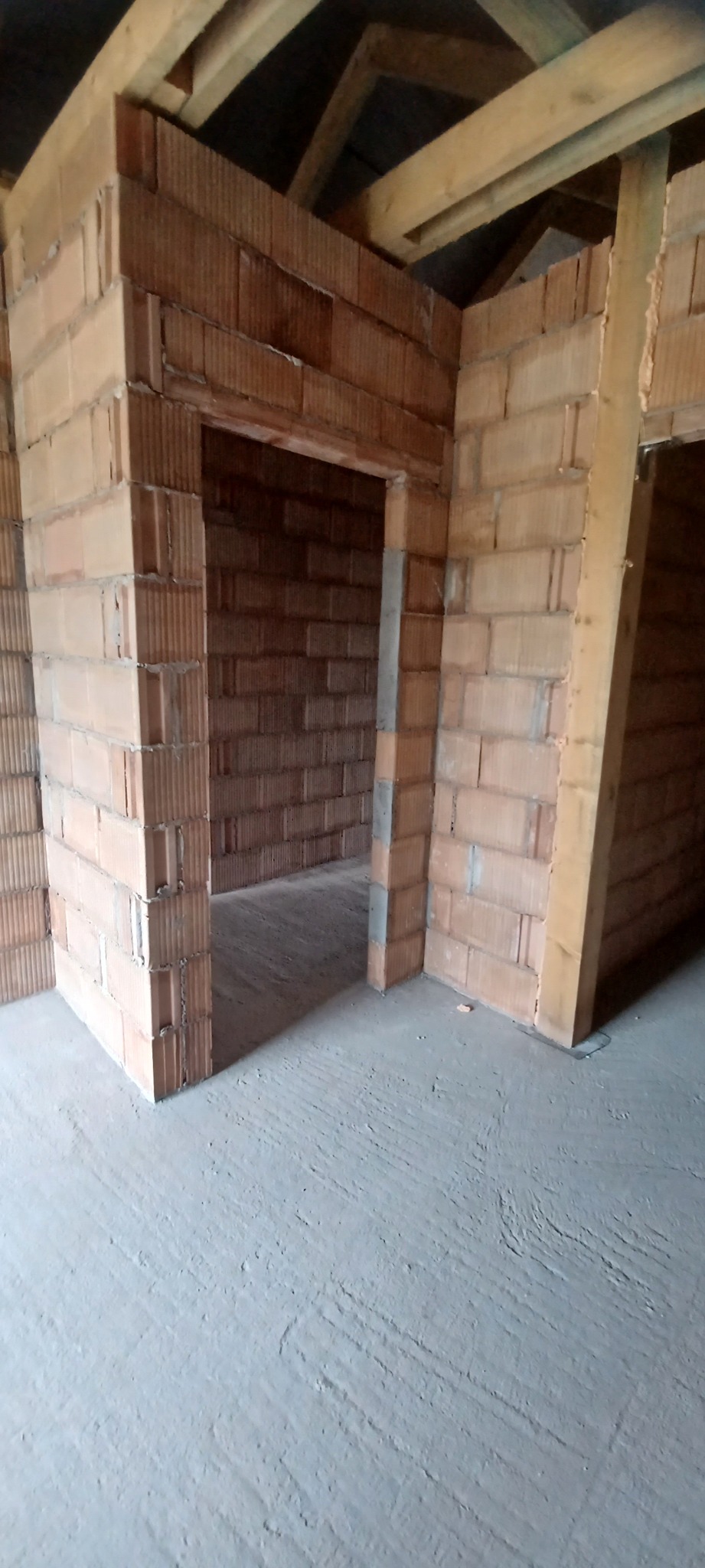 Wnętrze domu w trakcie budowy z widocznymi ścianami z czerwonej cegły dziurawki, drewnianą konstrukcją dachu i surową betonową podłogą.