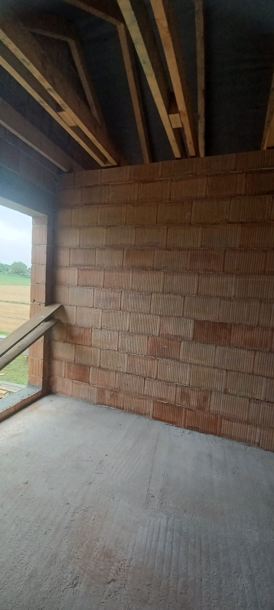Wnętrze surowego budynku z widocznymi ścianami z pustaków ceramicznych, drewnianą konstrukcją dachu i wylewką podłogową, z widokiem na pola przez otwór okienny.