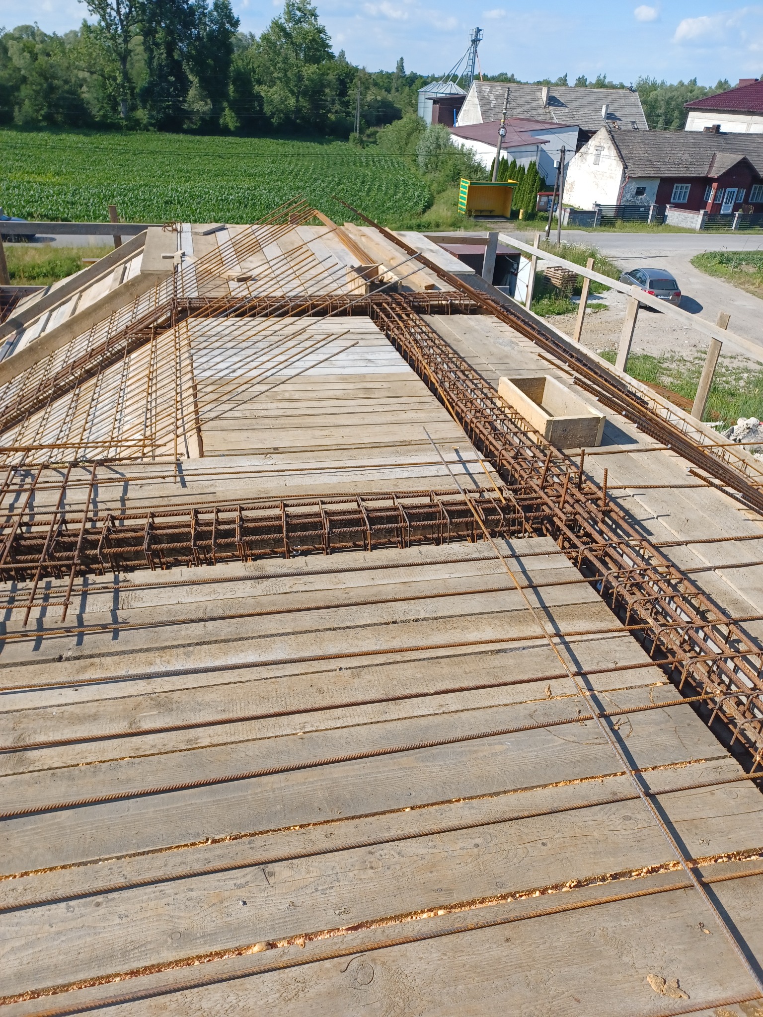 Zbrojenie stropu na drewnianym szalunku, widoczne pręty zbrojeniowe i betonowa skrzynka, w tle zabudowa wiejska i pole uprawne.