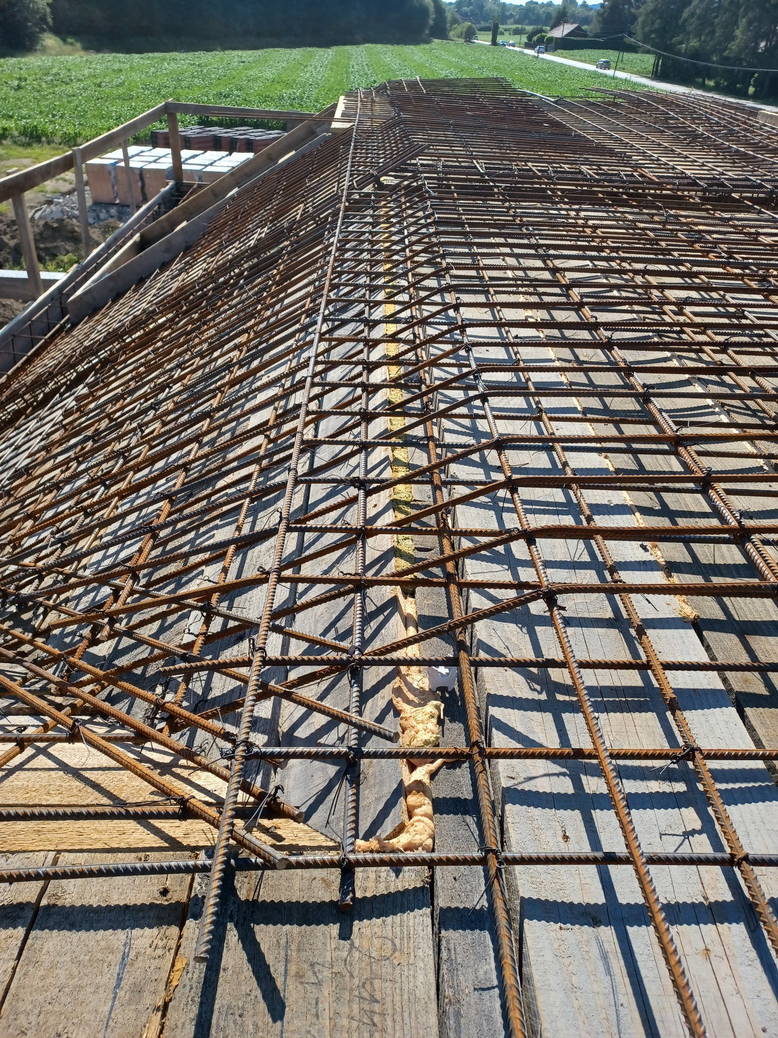 Zbrojenie na deskowaniu, widok z góry na konstrukcję przygotowaną do zalania betonem, w tle zielone pole i fragment konstrukcji drewnianej.