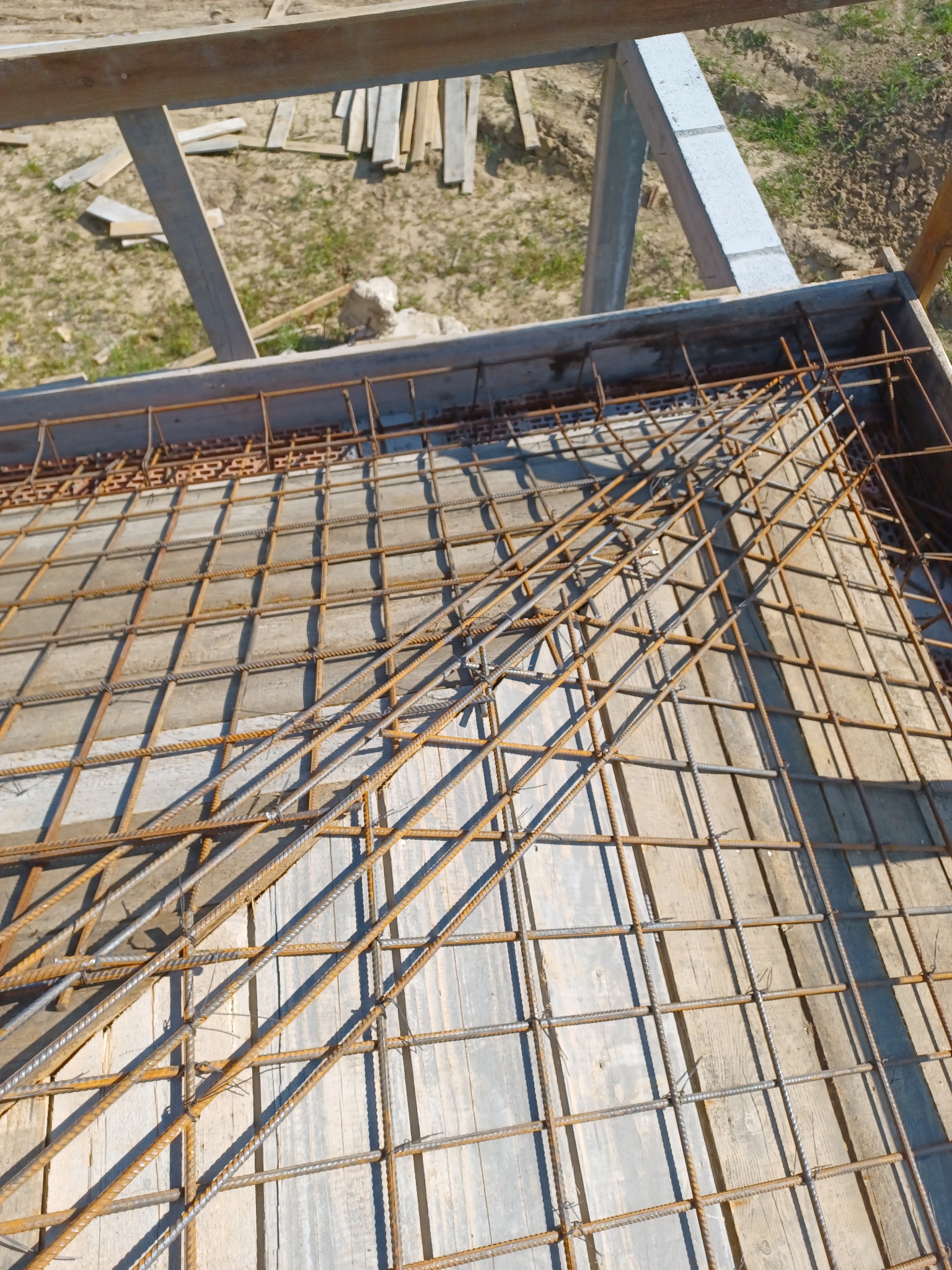 Zbrojenie fundamentu z siatki stalowej na deskowaniu, widoczne elementy konstrukcji szalunkowej i fragment gruntu w tle.