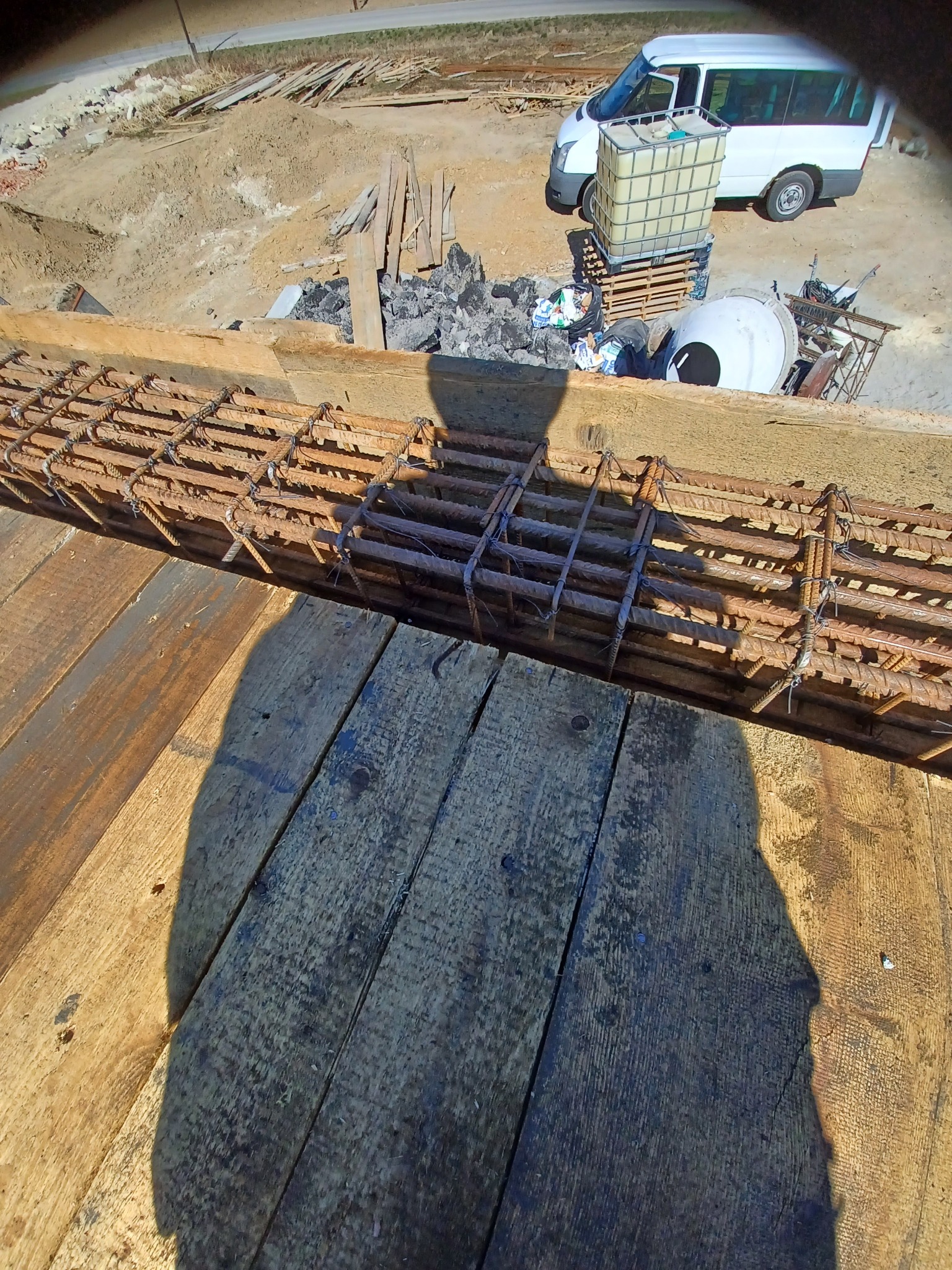 Zbrojenie fundamentu w trakcie montażu, widoczny szalunek z desek i cień osoby robiącej zdjęcie, w tle teren budowy z zaparkowanym busem i betoniarką.