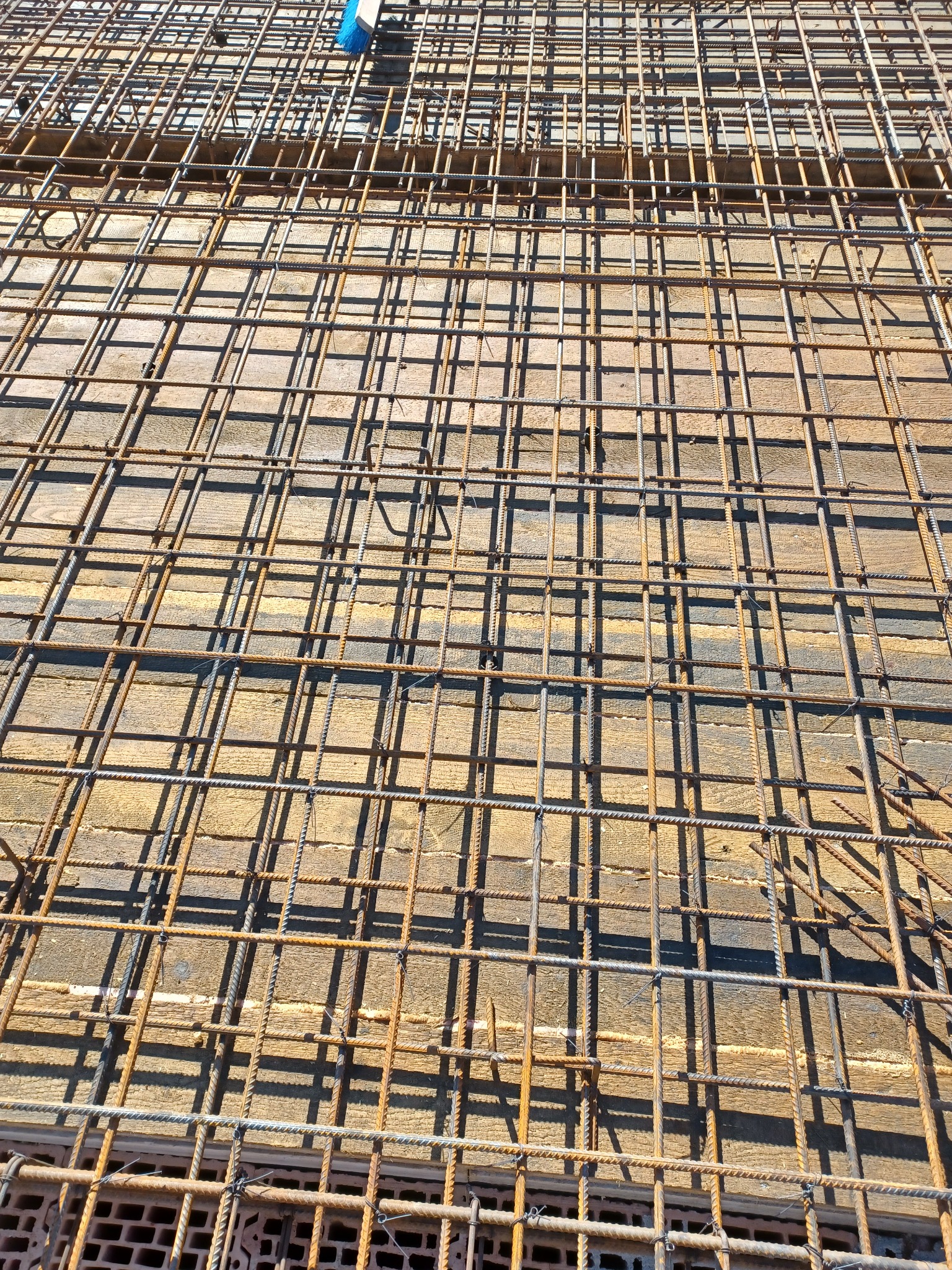 Siatka zbrojeniowa ułożona na deskowaniu, przygotowana do zalania betonem, widoczne cegły w tle, z góry widoczny fragment niebieskiej miotły.