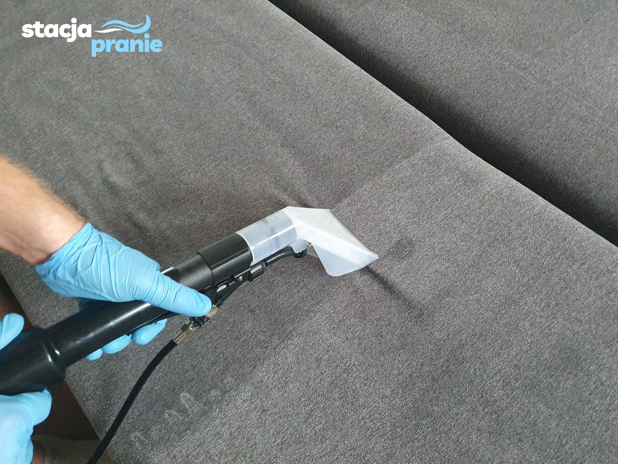 Czyszczenie szarej, tekstylnej kanapy za pomocą profesjonalnego odkurzacza ekstrakcyjnego, widoczne ręce w niebieskich rękawiczkach ochronnych oraz logo firmy w lewym górnym rogu.