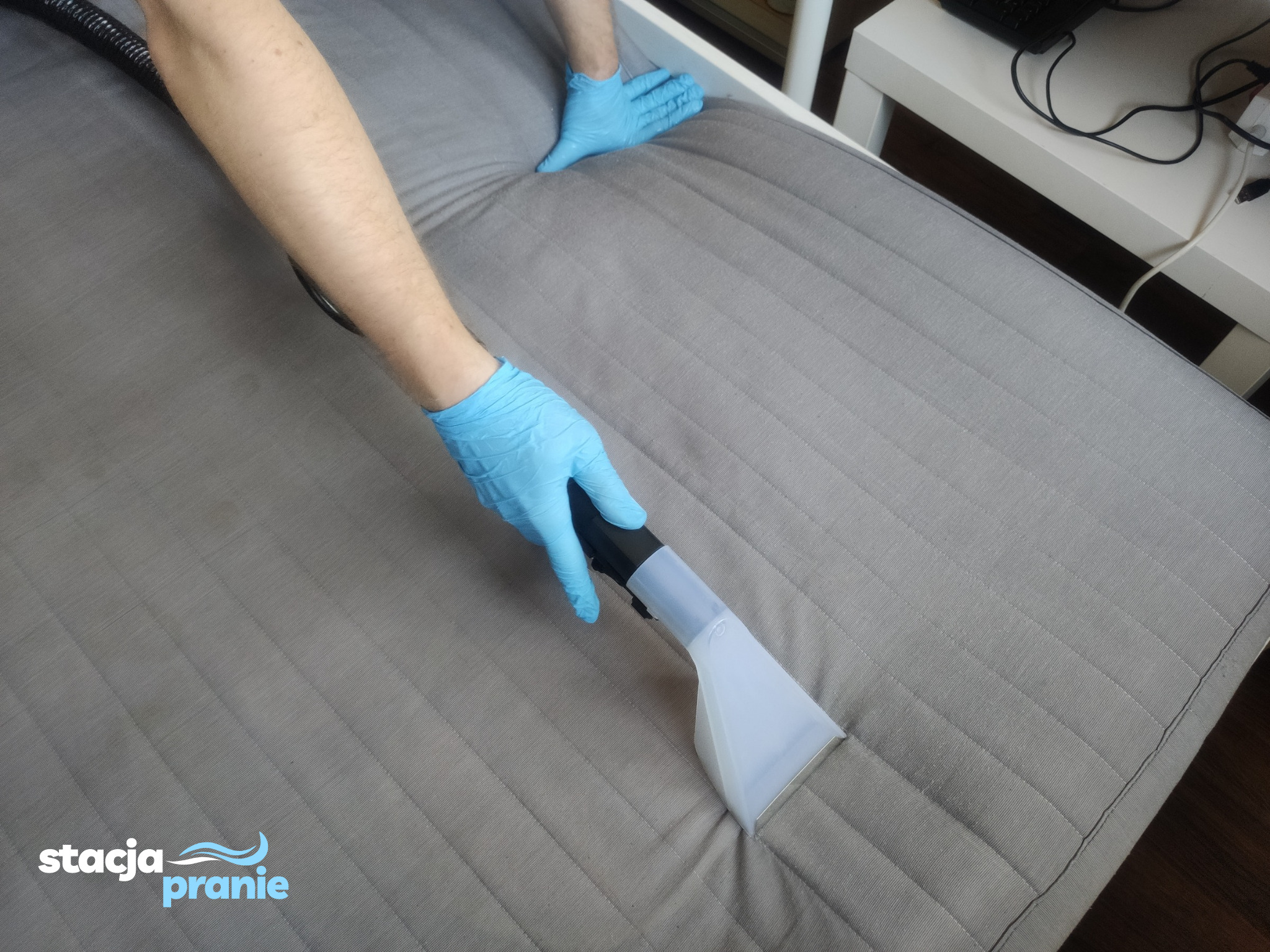 Czyszczenie jasnoszarego materaca na łóżku za pomocą ekstraktora do tapicerki, widoczne ręce w niebieskich rękawicach ochronnych.
