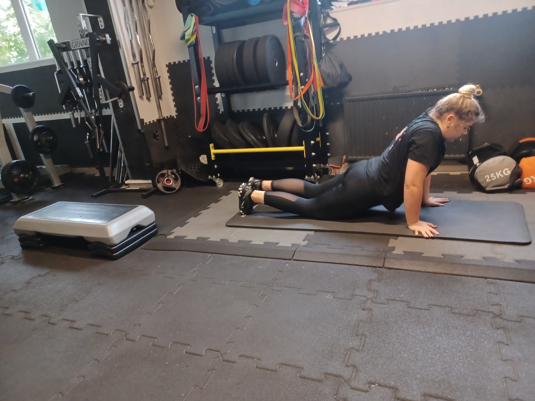 Kobieta w czarnym stroju sportowym wykonuje ćwiczenie rozciągające na macie w siłowni; w tle widoczne obciążenia i sprzęt do ćwiczeń.