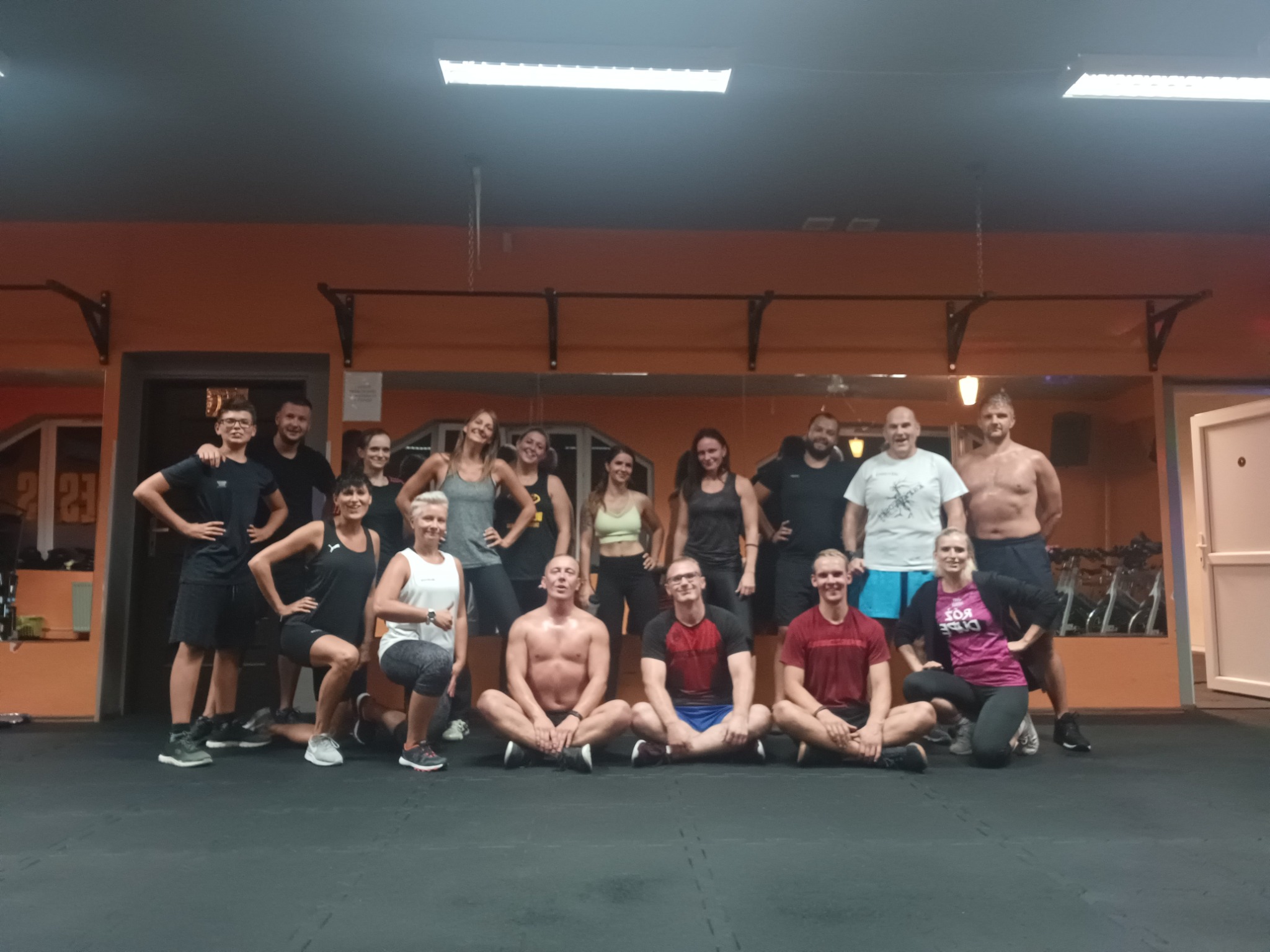 Grupa zadowolonych osób po treningu personalnym, część stoi, część siedzi na podłodze w siłowni z pomarańczowymi ścianami.