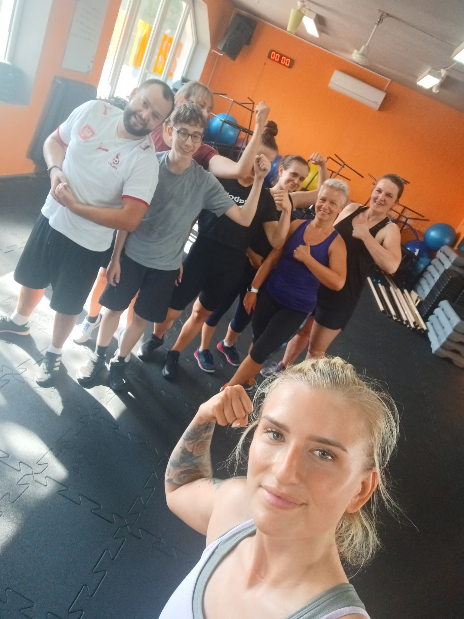 Grupa uśmiechniętych osób w sportowych strojach pozuje w sali fitness, z widocznymi hantlami i piłkami do ćwiczeń w tle, z blondynką z tatuażem na ramieniu na pierwszym planie.