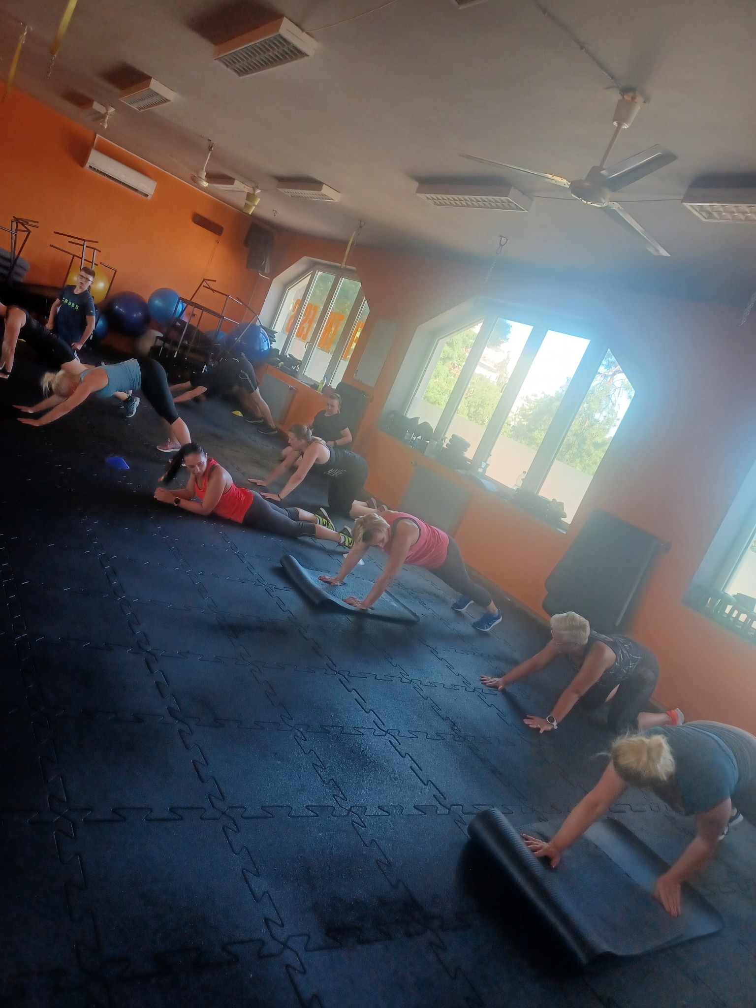Grupowe ćwiczenia w jasnej sali fitness, osoby wykonują ćwiczenia na macie, widoczne okna i sprzęt sportowy w tle.