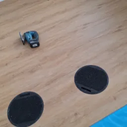 Układanie jasnych paneli podłogowych z okrągłymi otworami na kable, widoczna wyrzynarka akumulatorowa Makita, niebieska folia ochronna przy ścianie.