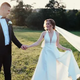 Uśmiechnięta para młoda trzymająca się za ręce na zielonej łące w promieniach zachodzącego słońca, z welonem powiewającym na wietrze.