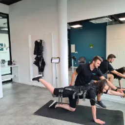 Kobieta w czarnym stroju EMS ćwiczy na macie pod nadzorem mężczyzny, widoczne odbicie w lustrze, sprzęt EMS na wieszaku w tle.