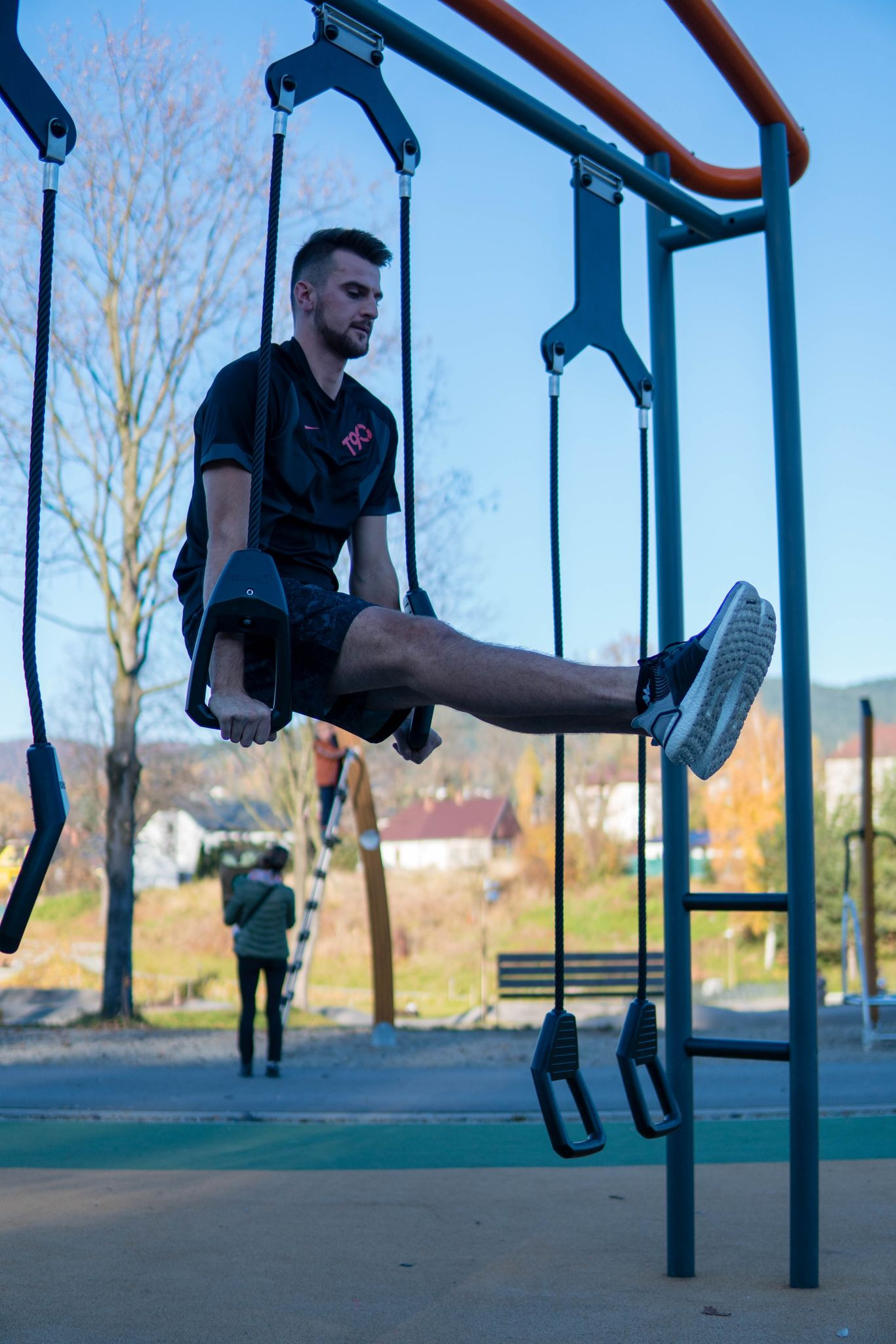 Mężczyzna wykonuje ćwiczenie na drążku gimnastycznym na świeżym powietrzu, unosząc nogi poziomo; w tle drzewa i budynki.