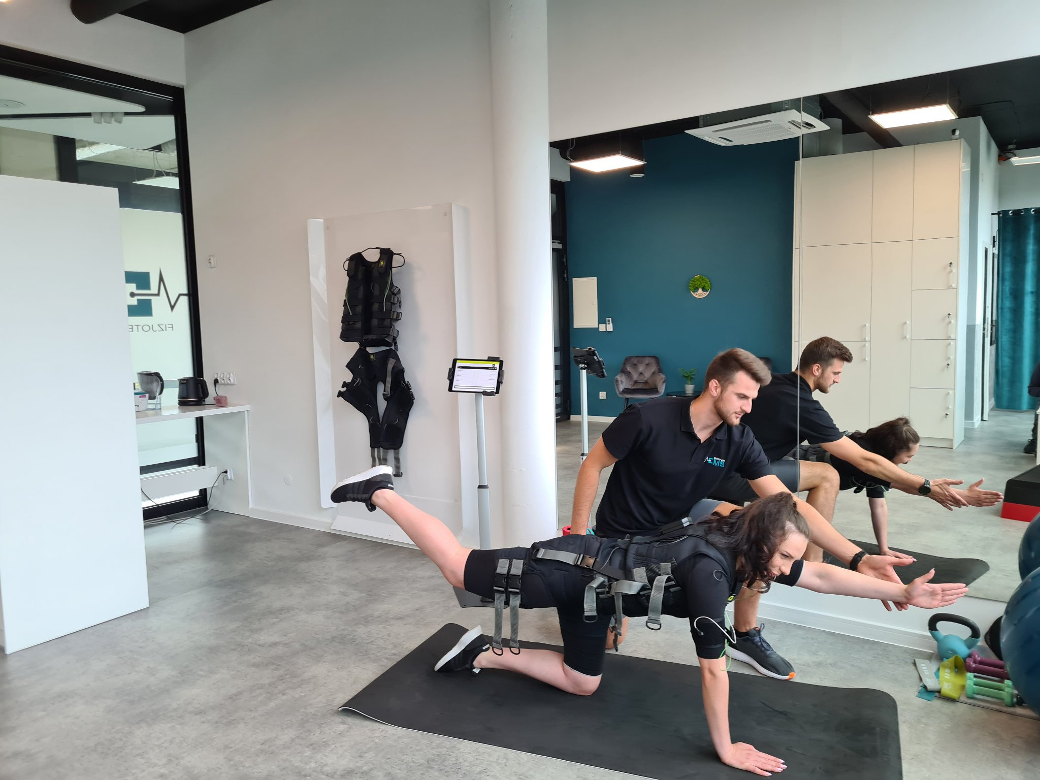 Kobieta w czarnym stroju EMS ćwiczy na macie pod nadzorem mężczyzny, widoczne odbicie w lustrze, sprzęt EMS na wieszaku w tle.