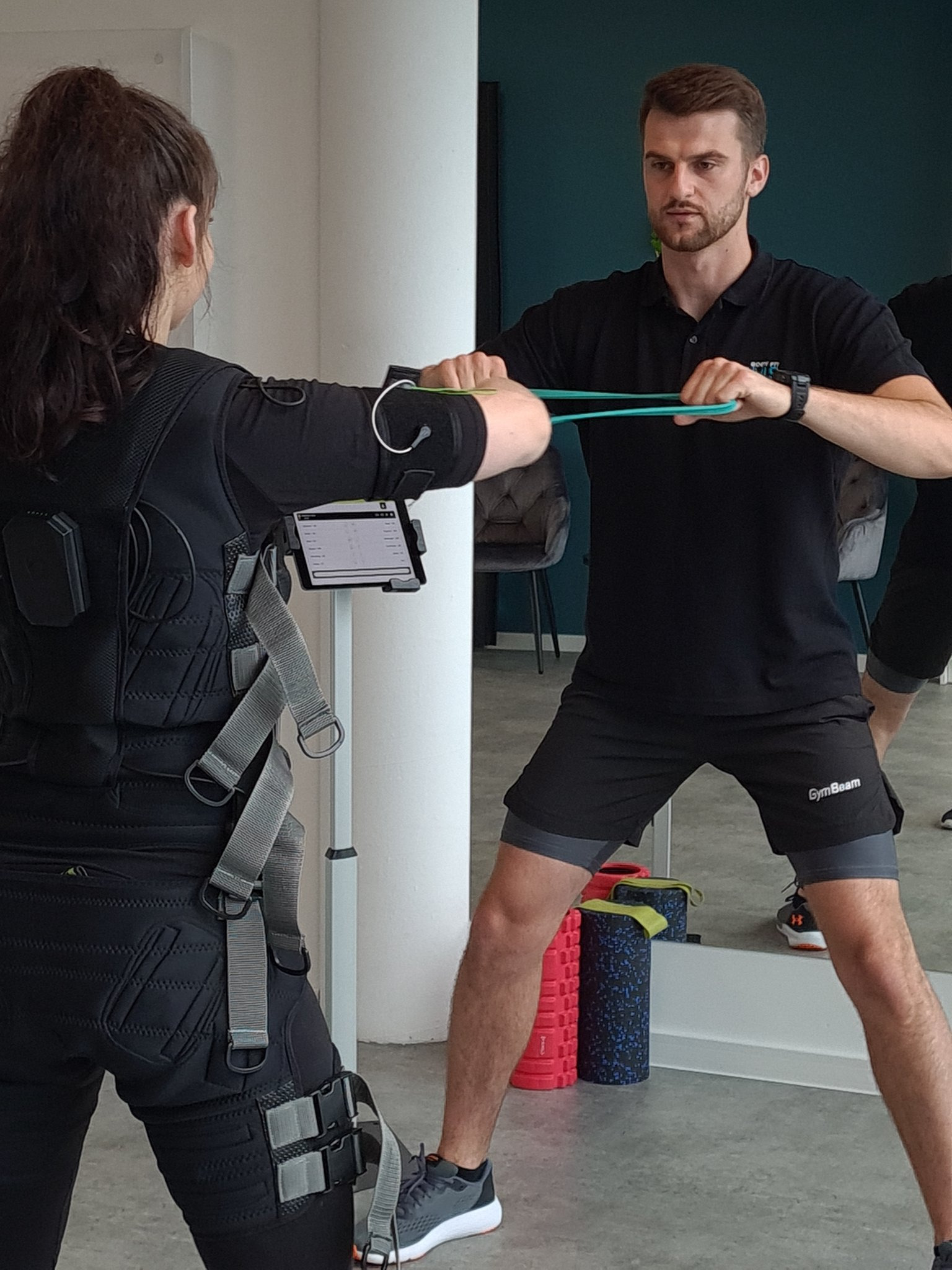 Kobieta w kombinezonie EMS ćwiczy z gumą pod okiem trenera, widoczny tablet z ustawieniami treningu, w tle lustro i wałki do masażu.