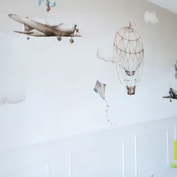 Dziecięcy pokój z dekoracyjną ścianą: akwarelowe malunki samolotów, balonu i latawca na białym tle z białą lamperią.