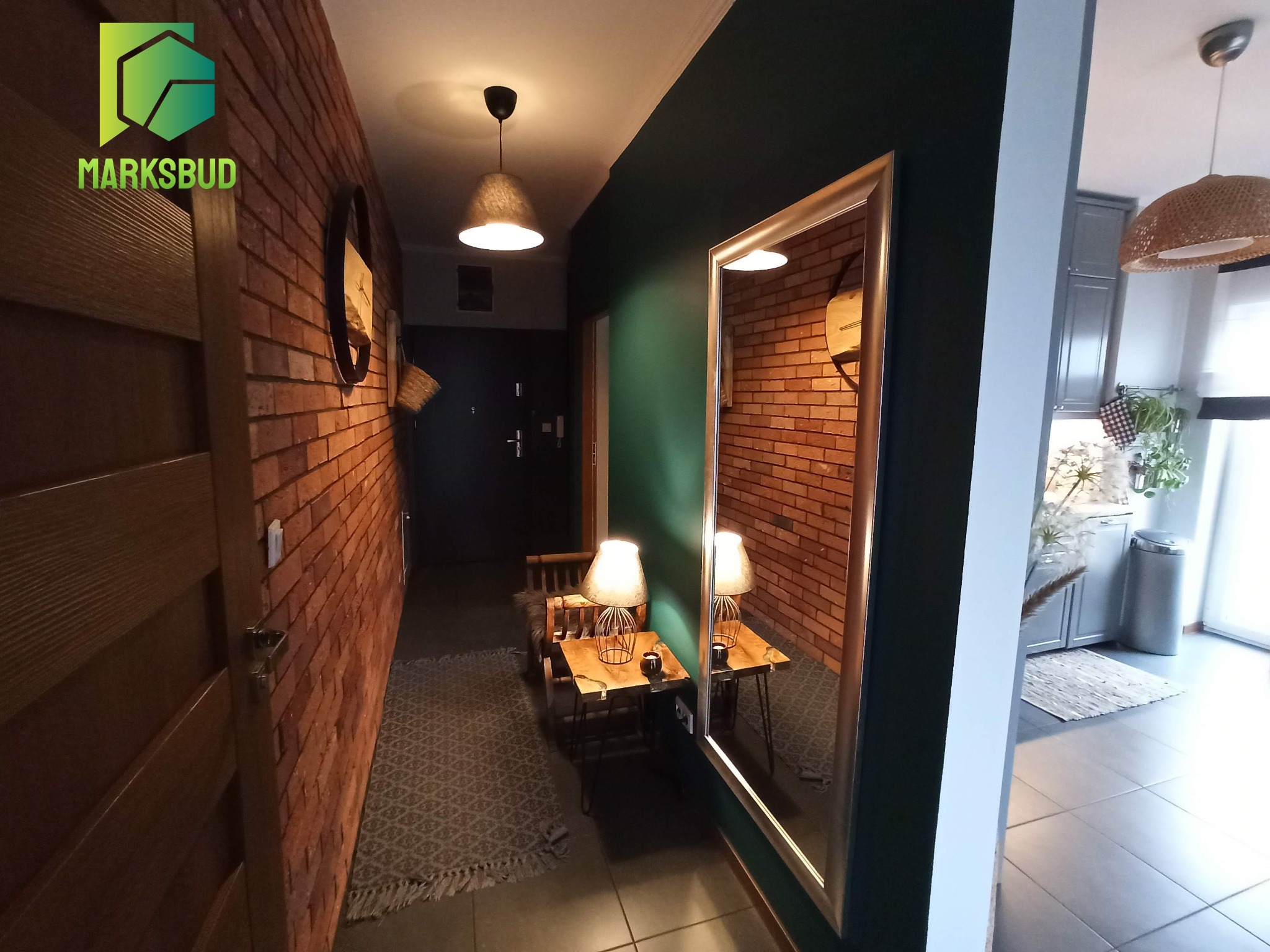 Korytarz z ceglaną ścianą, lustrem w srebrnej ramie, zieloną ścianą, drewnianymi drzwiami, lampą wiszącą i logo firmy MARKSBUD w lewym górnym rogu.