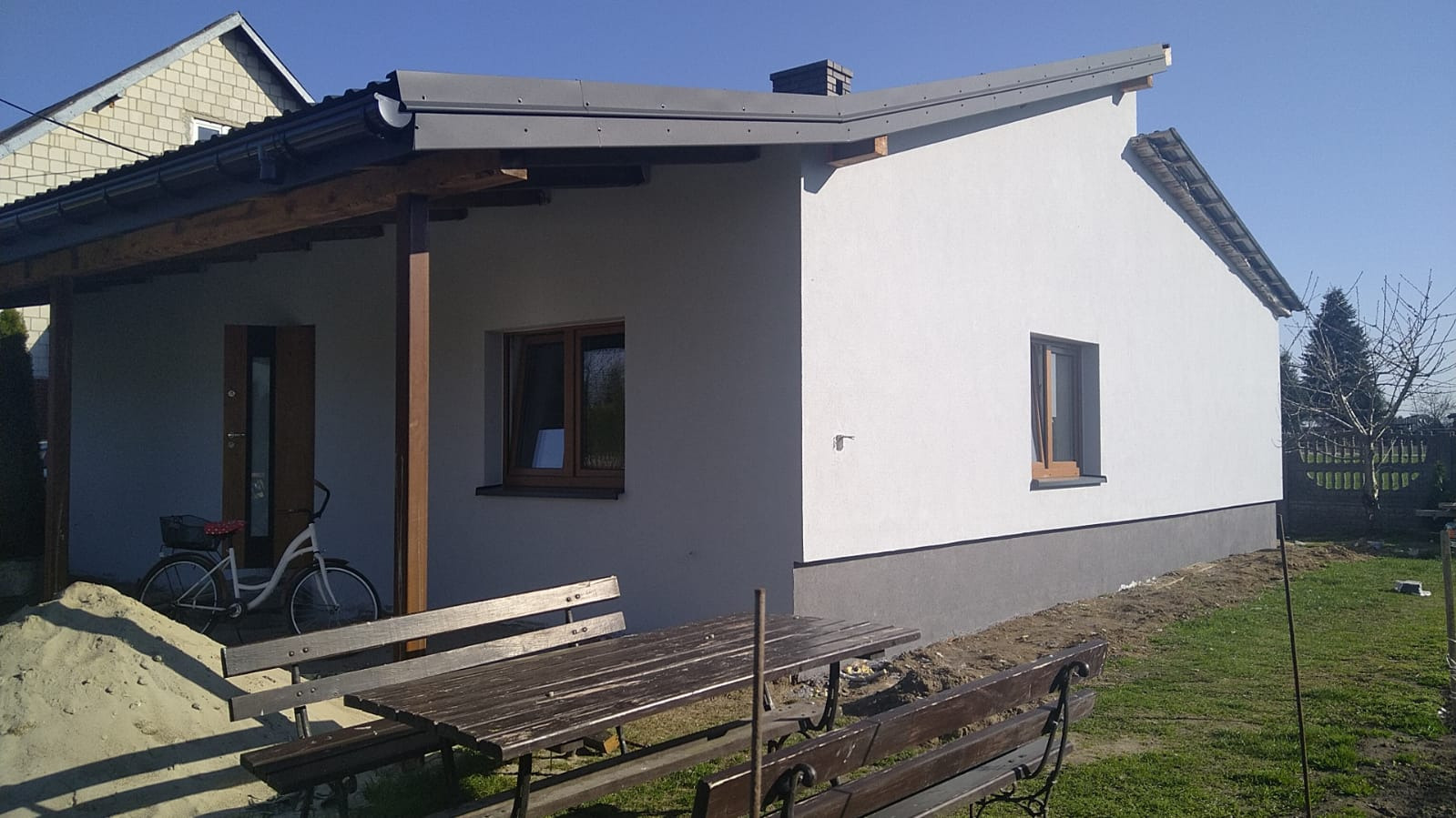 Otynkowany, parterowy dom z szarą podmurówką i drewnianą werandą. Na pierwszym planie stół piknikowy i rower oparty o ścianę. Błękitne niebo w tle.