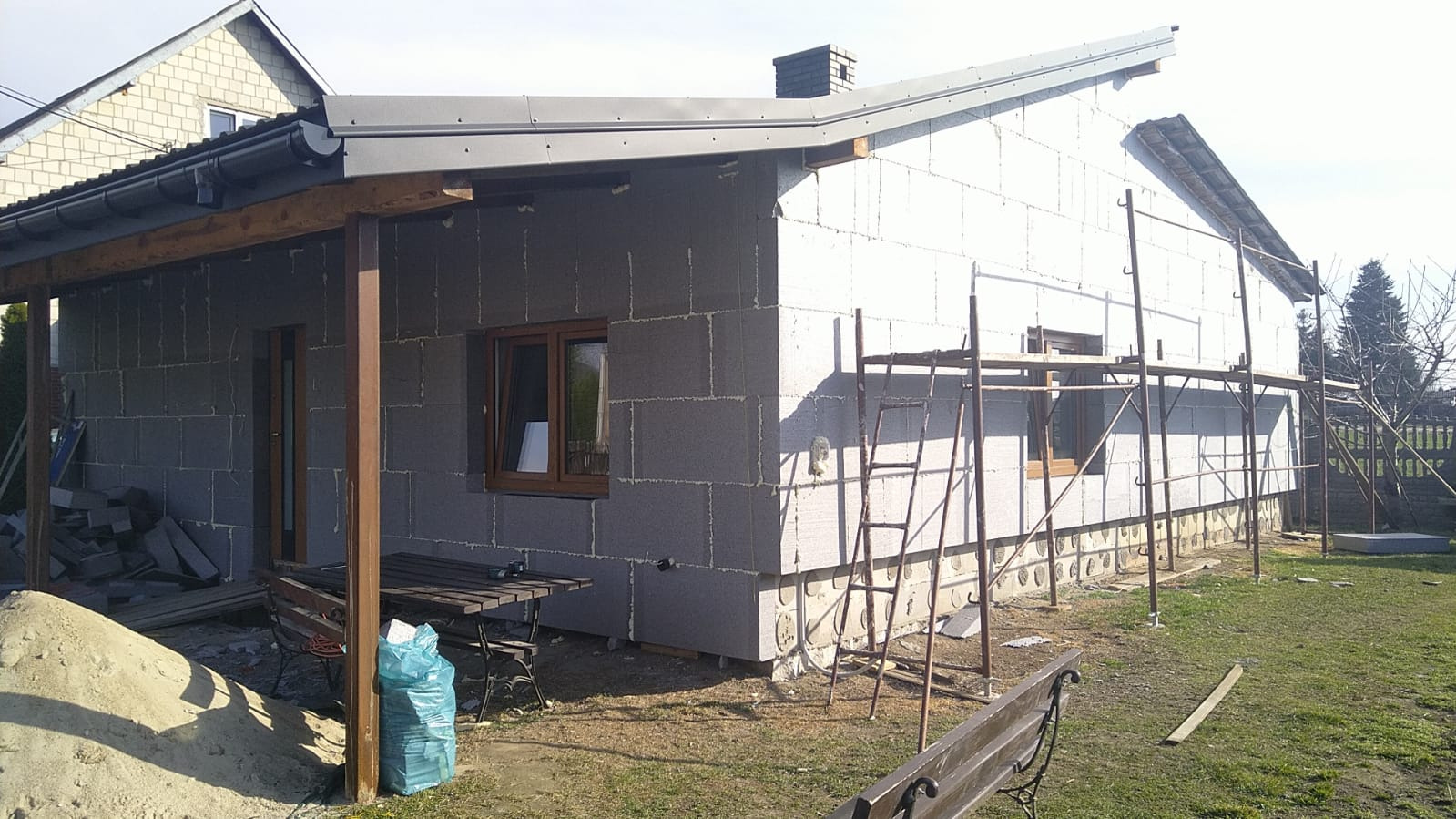 Dom w trakcie ocieplania: ściany pokryte styropianem, rusztowania, okna z brązowymi ramami. Widoczny fragment dachu i podwórka z elementami budowlanymi.