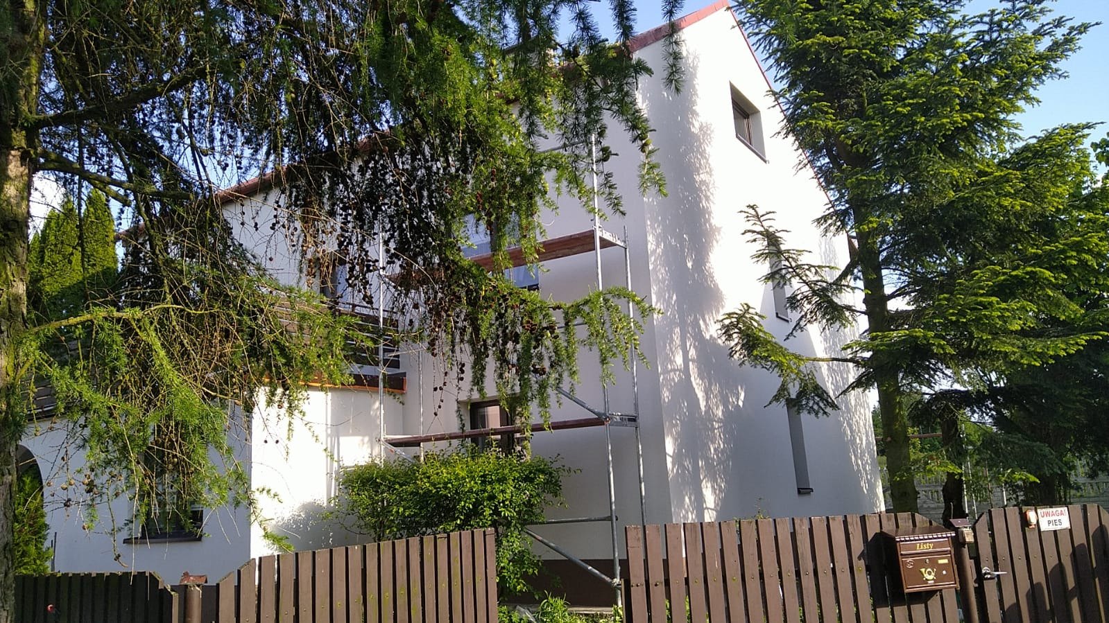 Biały dom jednorodzinny z rusztowaniem przy ścianie, częściowo zasłonięty drzewami i brązowym ogrodzeniem. Widoczna skrzynka na listy.