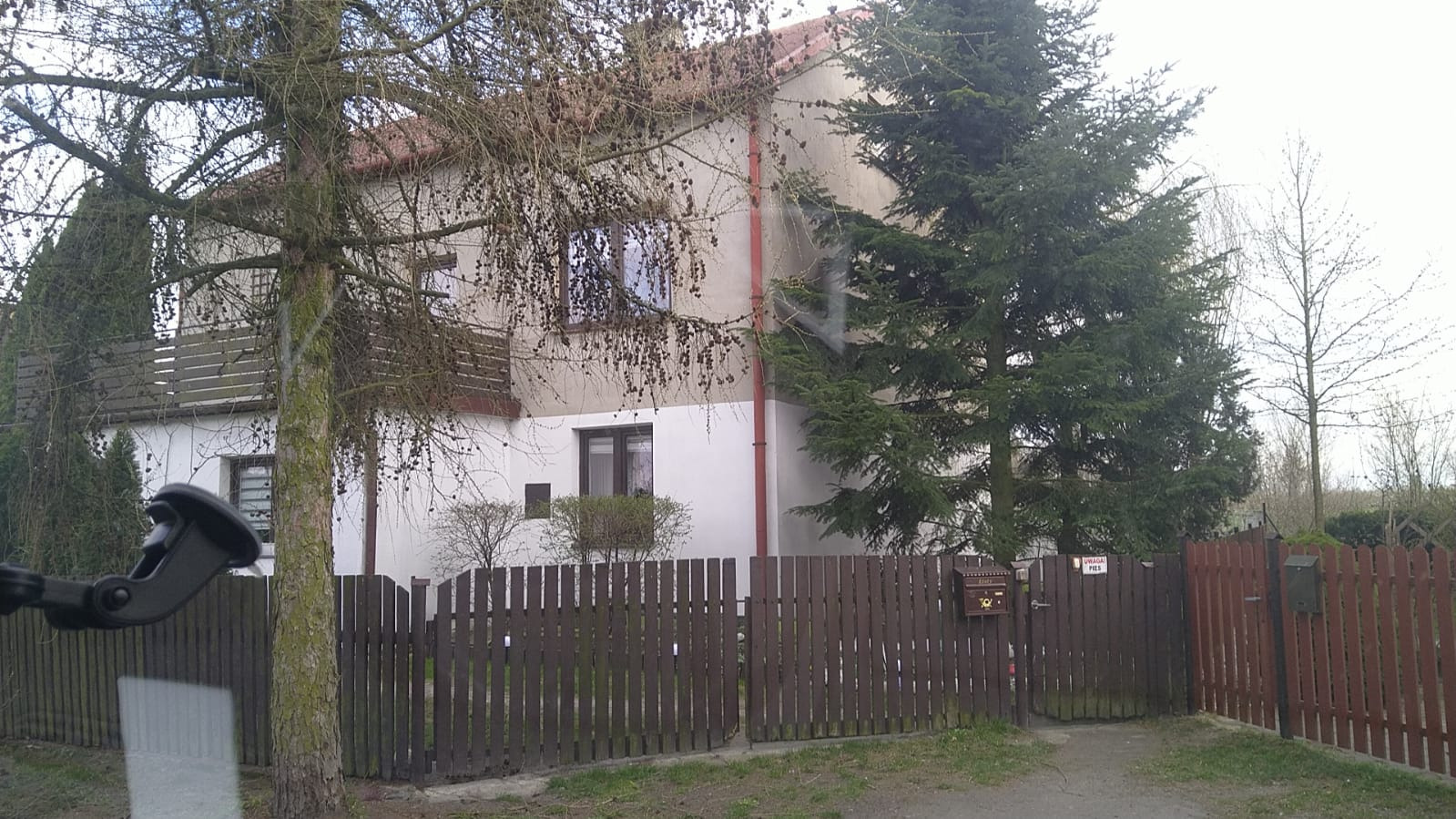 Dwukondygnacyjny dom z balkonem, brązową stolarką okienną i ogrodzeniem z bramką, otoczony zielenią drzew. Widoczna skrzynka pocztowa i tabliczka.