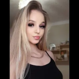 Portret młodej kobiety z długimi blond włosami, mocnym makijażem oczu i błyszczącymi ustami, pozującej na rozmytym tle wnętrza.