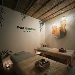 Dwa łóżka do tajskiego i balijskiego zabiegu, udekorowane koszykami z akcesoriami, oświetlone lampami z frędzlami, z logo Thai Organic na ścianie i motywami liści palmowych.