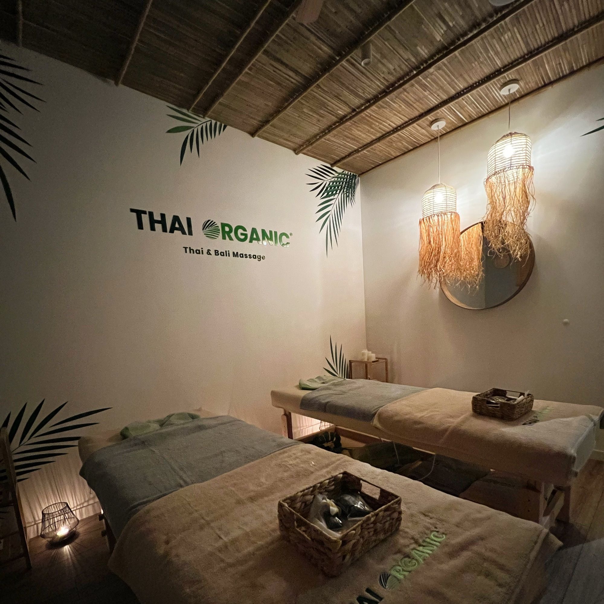Dwa łóżka do tajskiego i balijskiego zabiegu, udekorowane koszykami z akcesoriami, oświetlone lampami z frędzlami, z logo Thai Organic na ścianie i motywami liści palmowych.