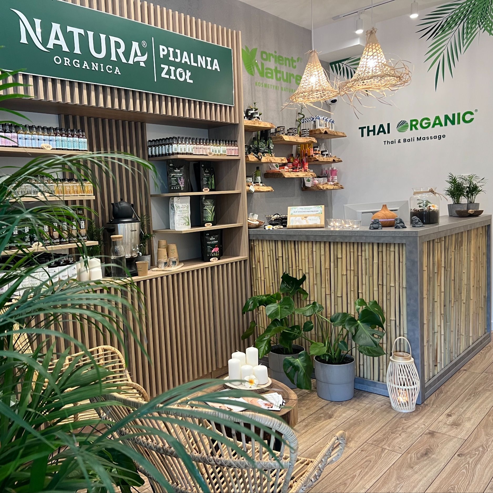 Wnętrze salonu z ziołami i kosmetykami naturalnymi, widoczne półki z produktami, lada recepcyjna z bambusa, rattanowe lampy i fotele, oraz rośliny doniczkowe, na ścianie logo Thai Organic Thai &...