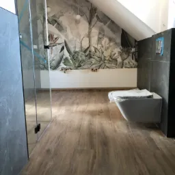 Łazienka w trakcie wykańczania, z zamontowaną toaletą w folii ochronnej, szklaną kabiną prysznicową i fototapetą z motywem roślinnym na ścianie pod skosem poddasza.