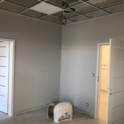 Ekipa - Wnętrze po malowaniu ścian na szaro we Wrocławiu, z widocznym sufitem podwieszanym, dwoma białymi drzwiami z poziomymi frezami oraz sprzętem malarskim przy ścianie.