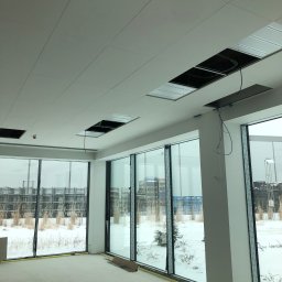 Ekipa - Wnętrze biura w stanie deweloperskim z dużymi oknami, widoczne niedokończone sufity podwieszane z wystającymi kablami, surowa podłoga i zimowy krajobraz za oknami.