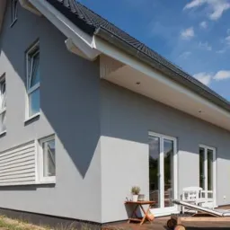 Szary dom jednorodzinny z białymi oknami i drzwiami, widok z zewnątrz na elewację z fragmentem tarasu i meblami ogrodowymi w słoneczny dzień.