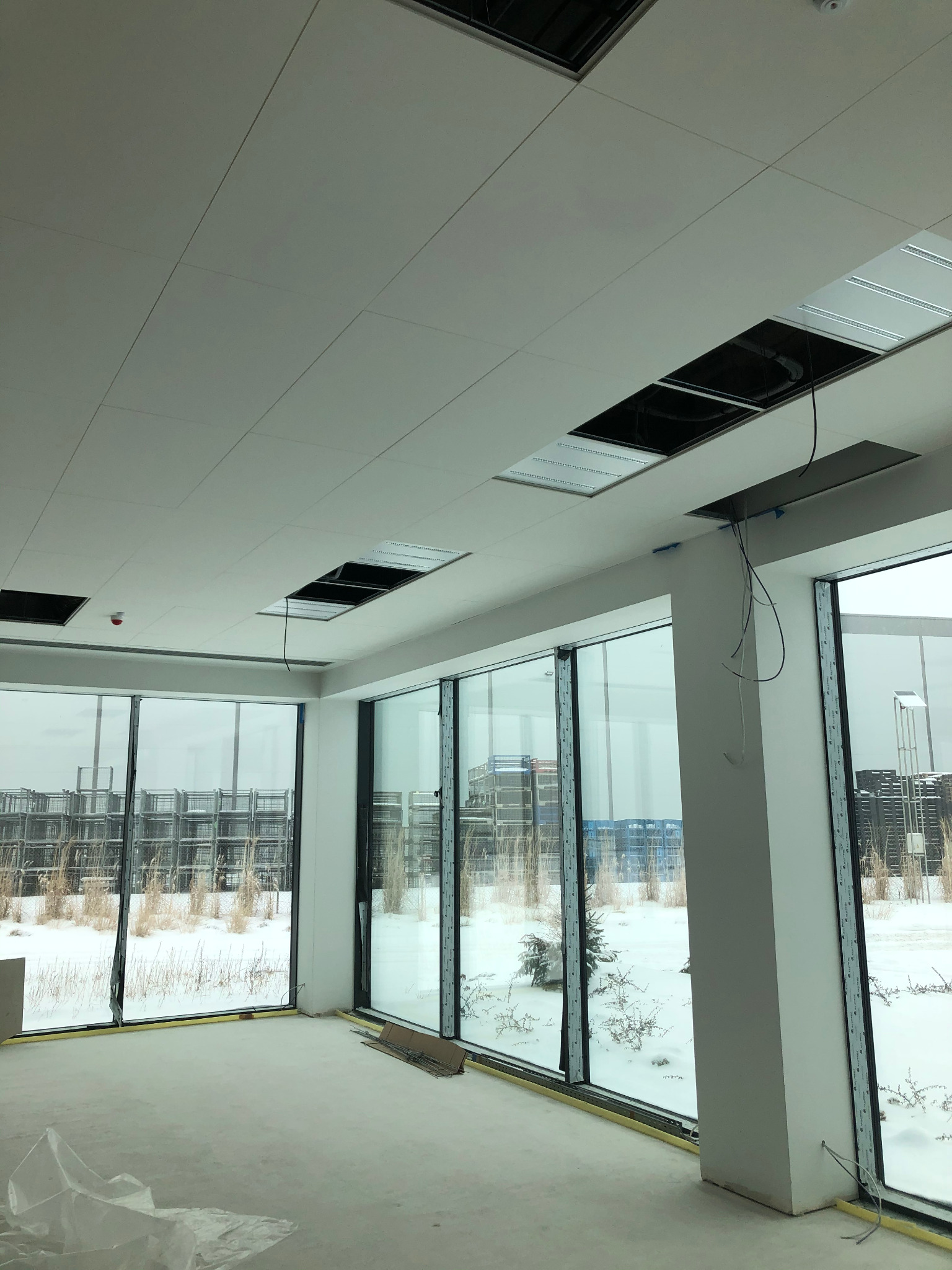 Wnętrze biura w stanie deweloperskim z dużymi oknami, widoczne niedokończone sufity podwieszane z wystającymi kablami, surowa podłoga i zimowy krajobraz za oknami.