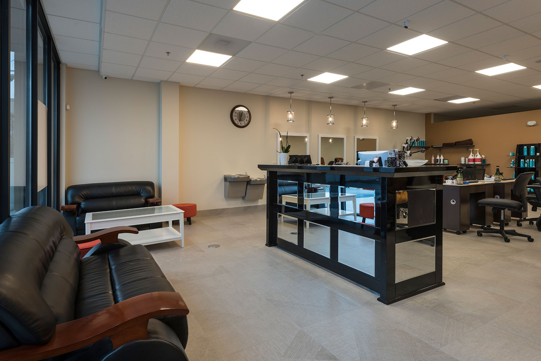 Przestronne wnętrze salonu kosmetycznego z recepcją, poczekalnią z czarnymi skórzanymi sofami i stanowiskami do manicure z lustrami i oświetleniem punktowym.