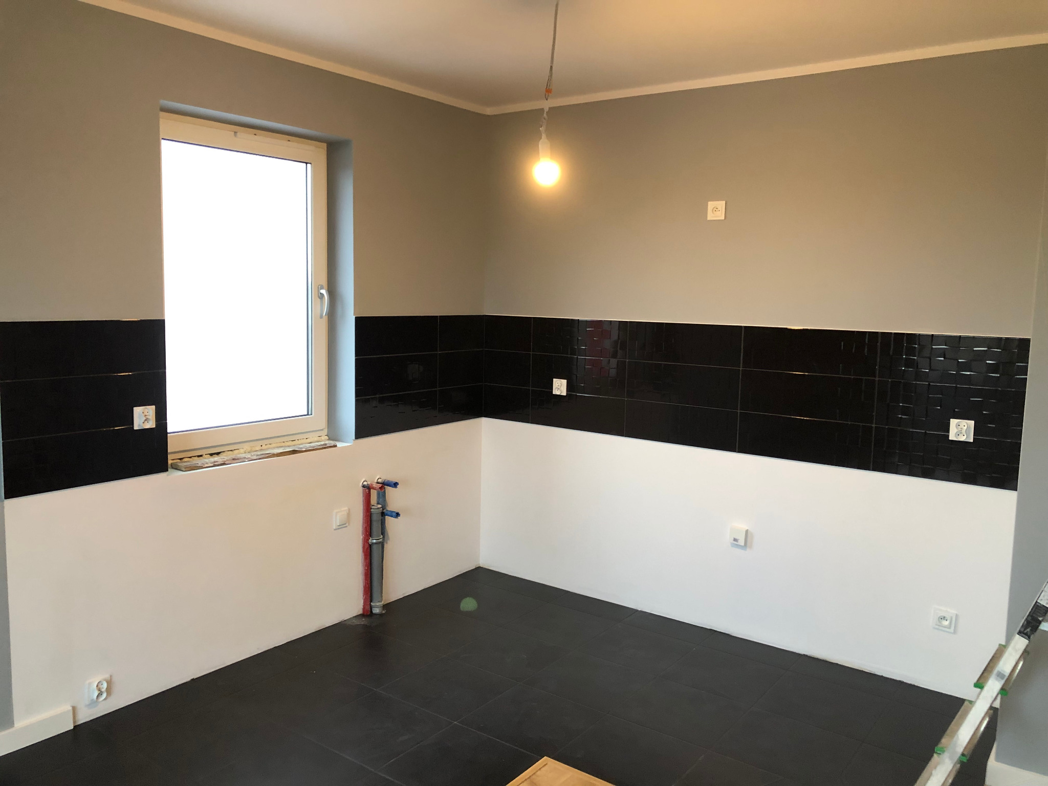Wnętrze pomieszczenia w trakcie prac wykończeniowych: szare ściany, czarne płytki do połowy wysokości, biała dolna część ścian, czarna podłoga z płytek, widoczne rury instalacyjne i drabina malarska.