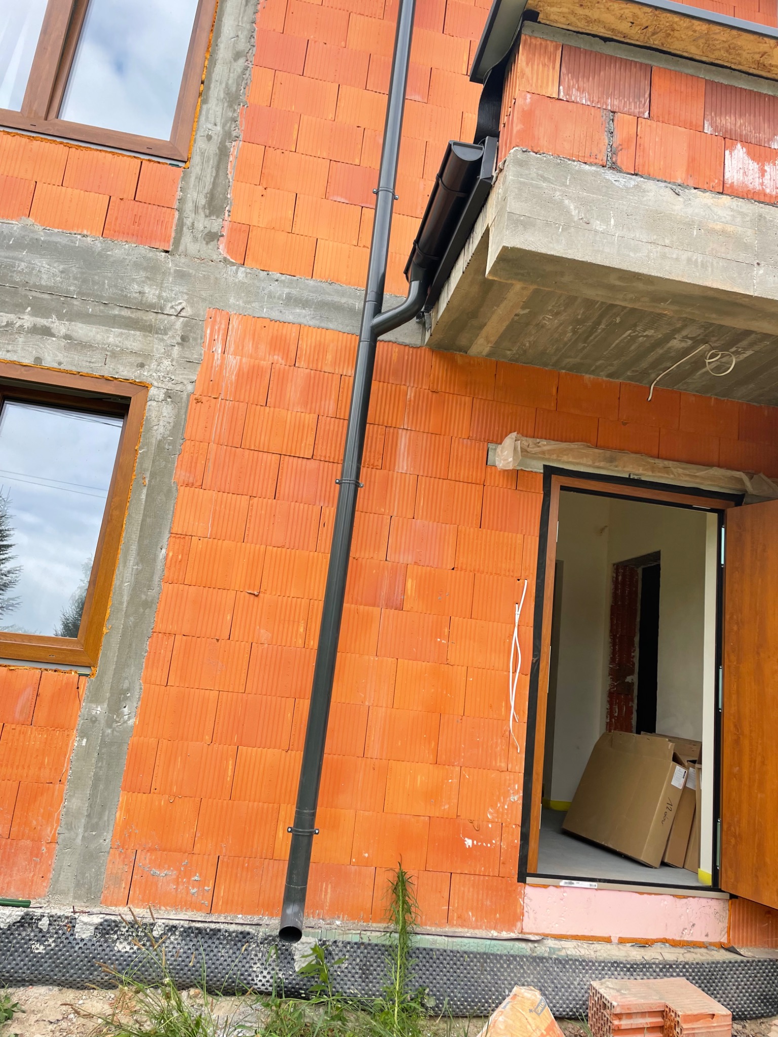 Surowy stan budynku z pomarańczowej cegły z zamontowaną czarną rynną spustową, widoczne okna z brązowymi ramami i otwarte drzwi wejściowe z kartonami w środku.