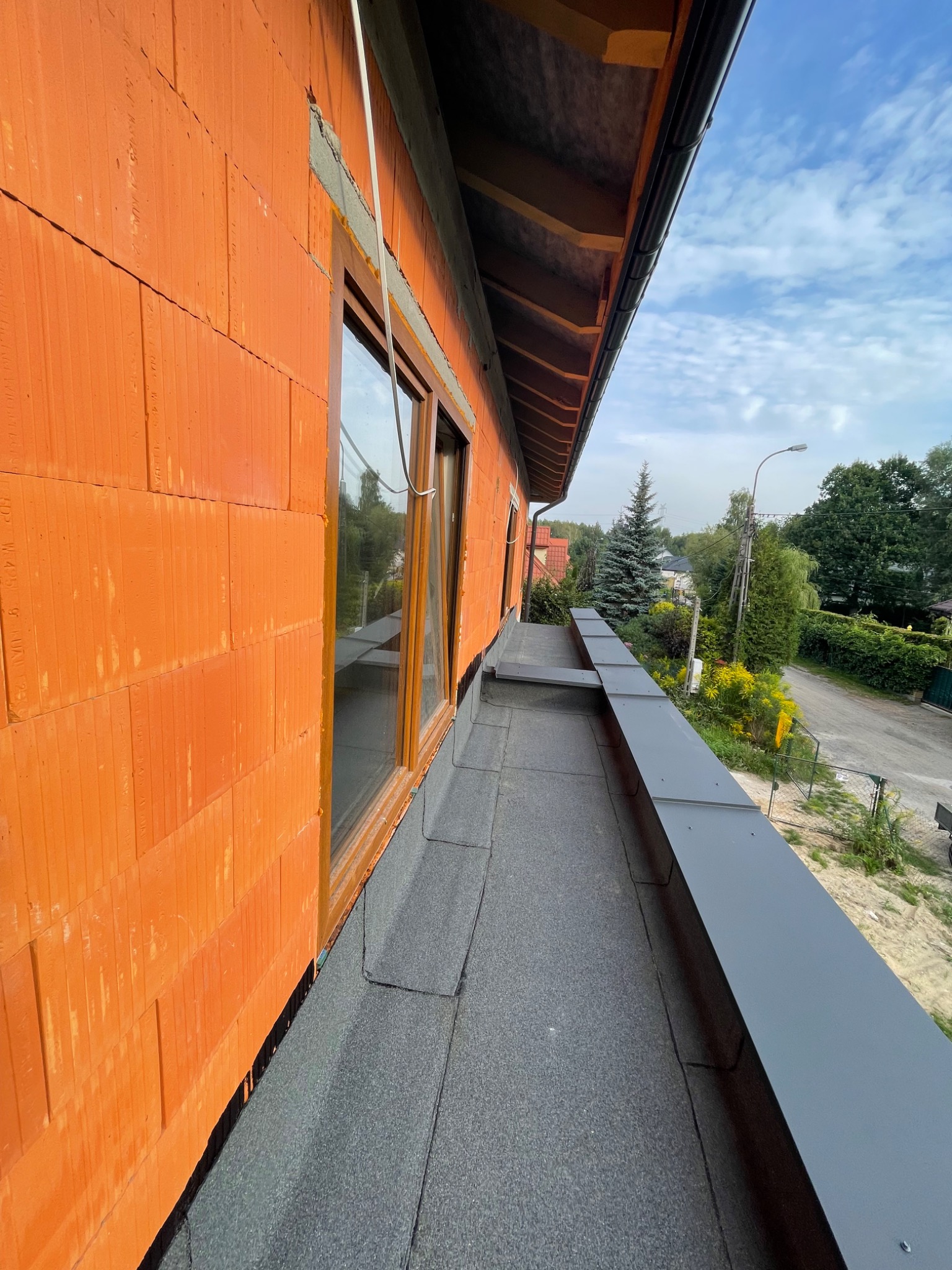 Surowy stan budynku z pomarańczowej cegły z zamontowanymi oknami, widoczne czarne pokrycie dachu i szare elementy wykończeniowe, w tle zabudowa jednorodzinna i zieleń.