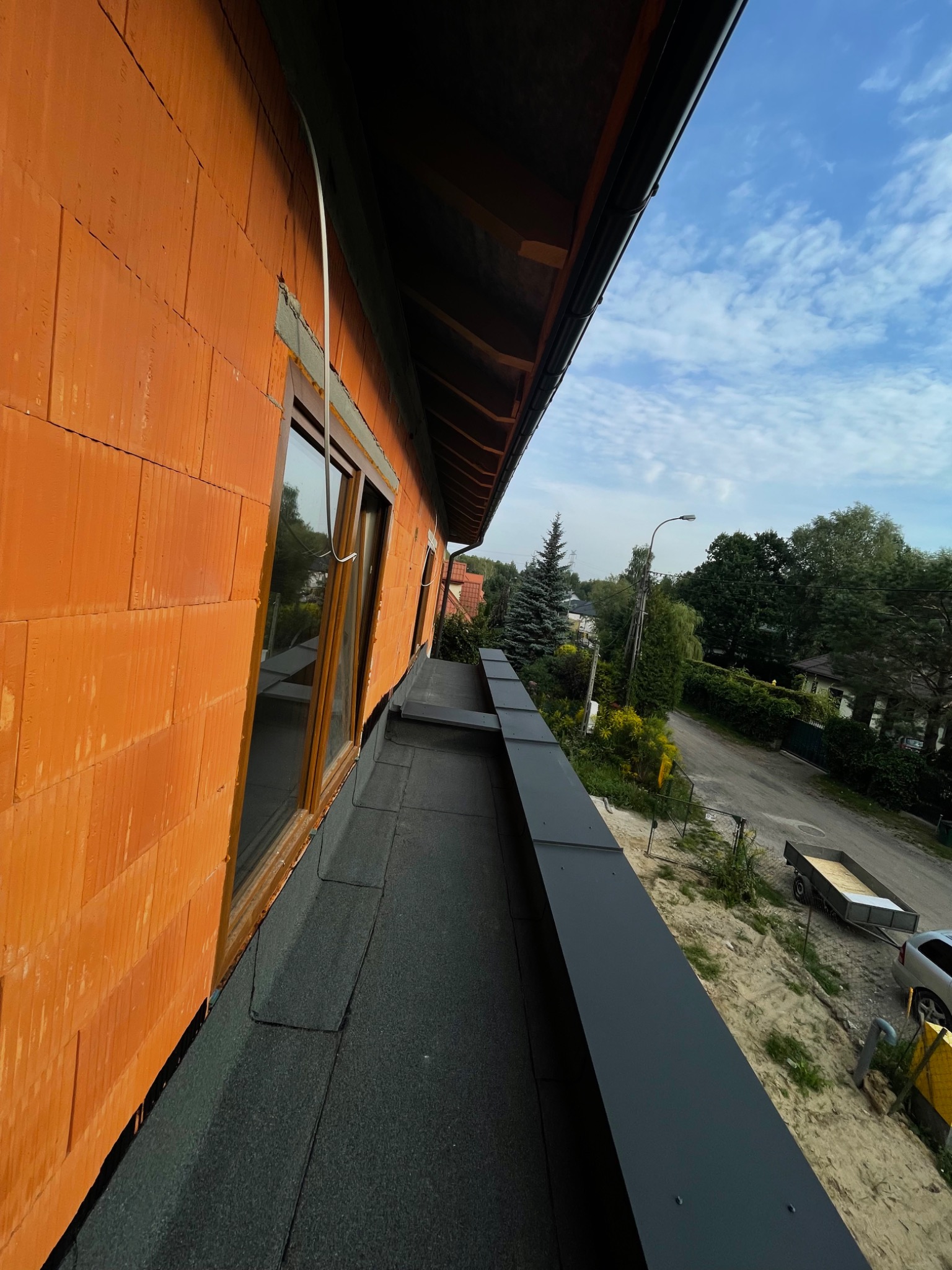 Nowa, czarna rynna zamontowana na dachu budynku z pomarańczowej cegły, widok z góry na okolicę.