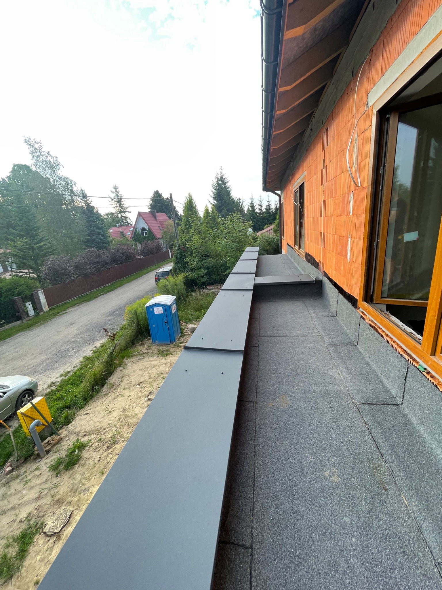 Szare, metalowe elementy wykończeniowe dachu płaskiego, widok z góry na nowo budowany dom z cegły ceramicznej, w tle ulica i domy jednorodzinne.